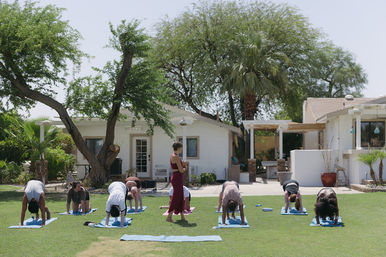Outdoor group yoga class on a sunny residential lawn, students on blue mats doing forward folds while an instructor in a red outfit walks among palm and shade trees in a backyard setting.