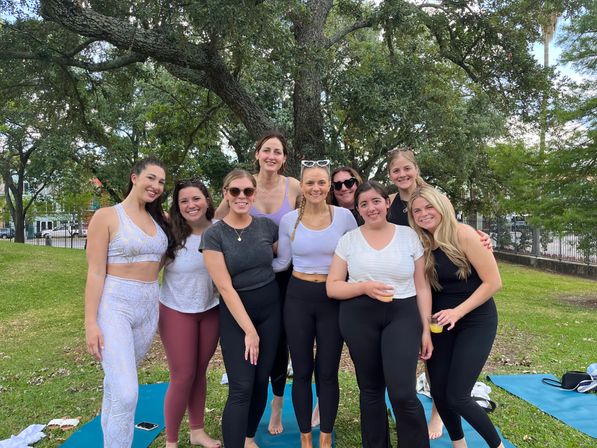 Group of nine women in activewear smiling on yoga mats under a large oak tree in a sunny park — an outdoor yoga gathering with some holding drinks.