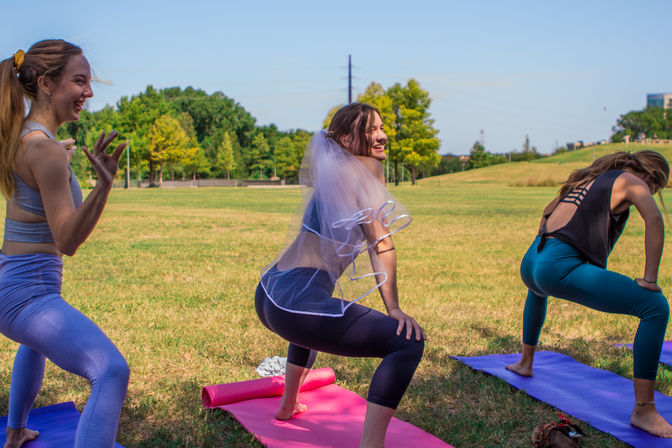 Three women practicing yoga squats on colorful mats in a sunny grassy park, the center participant laughing while wearing a white bachelorette veil — outdoor group yoga session.