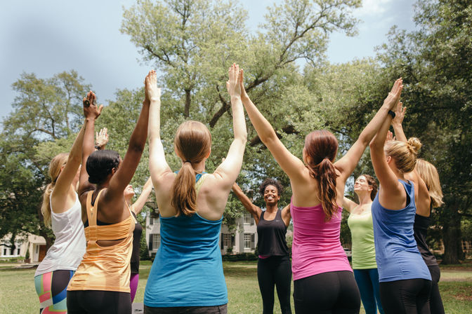 Women in colorful workout clothes standing in a circle on a sunny park lawn, arms raised, following a smiling instructor in an outdoor yoga/fitness class.