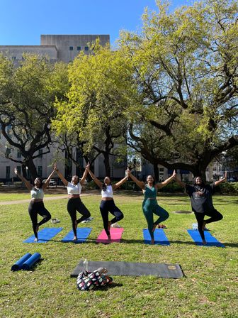 Five people balancing in tree pose during an outdoor yoga class on colorful mats on a sunny downtown park lawn beneath large leafy trees, with a stone building visible in the background.