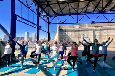 Group of people practicing tree pose on blue mats under a metal waterfront pavilion, with a sunlit city skyline visible across the water