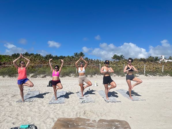 Five people practicing tree pose on yoga mats on a sunny sandy beach with palm trees and a bright blue sky — outdoor group yoga and beach fitness scene.