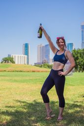 Woman in navy workout set and flip-flops holding a champagne bottle aloft in a sunny urban park, green lawn and city skyline of high-rise buildings in the background — outdoor fitness and celebration vibe.