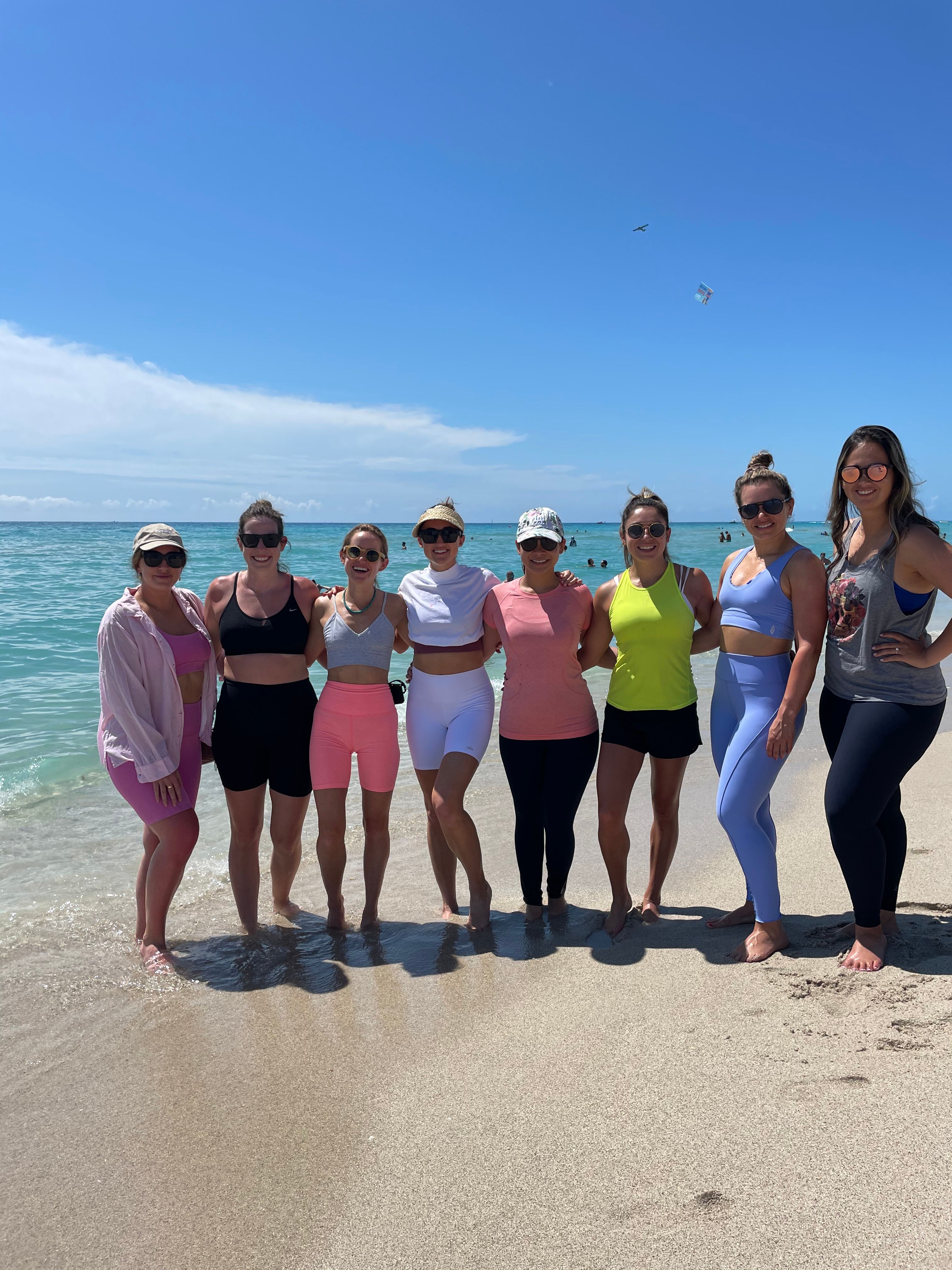 Eight women in colorful activewear standing barefoot at the water’s edge on a sunny turquoise beach, arms around each other with a clear blue sky and a kite overhead.
