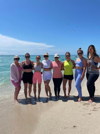 Eight women in colorful activewear standing barefoot at the water’s edge on a sunny turquoise beach, arms around each other with a clear blue sky and a kite overhead.