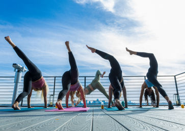 Five people doing leg-lift yoga poses on colorful mats during a sun-soaked rooftop yoga session with a city skyline and bright blue sky.