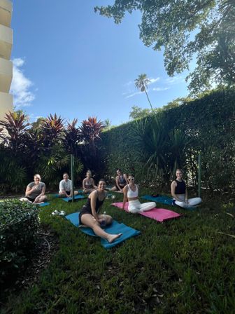 Group of seven people on colorful yoga mats in a sunny tropical garden courtyard with palm trees and blue sky, relaxed outdoor yoga session