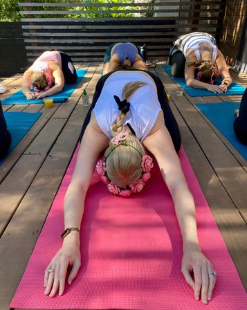 Group of women wearing flower crowns practicing child's pose on colorful yoga mats during an outdoor yoga class on a sunny wooden deck with drinks and greenery nearby.