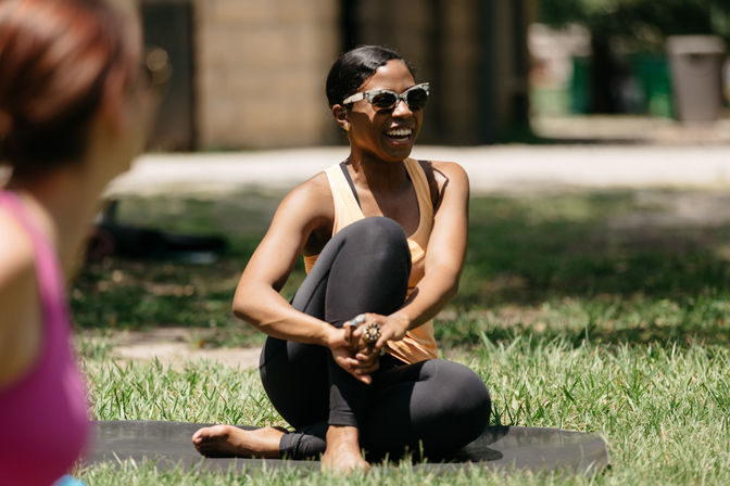 Smiling woman in sunglasses sitting cross-legged on a yoga mat in a sunny park during an outdoor yoga session, laughing and stretching.