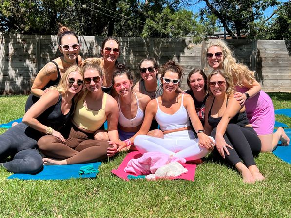 Outdoor yoga group of smiling women in activewear and sunglasses sitting together on colorful mats on a sunny backyard lawn with a wooden fence and trees