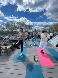 Rooftop outdoor yoga class on a wooden deck under a bright blue sky with puffy clouds, a small group balancing in tree pose on colorful mats with water bottles and benches nearby.