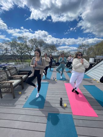 Rooftop outdoor yoga class on a wooden deck under a bright blue sky with puffy clouds, a small group balancing in tree pose on colorful mats with water bottles and benches nearby.