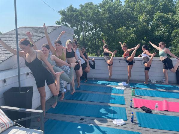 Sunny rooftop yoga class: a line of women balancing in dancer/standing bow pose on blue mats along a deck railing with green trees beyond.