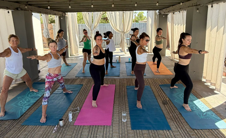 Mixed-age group of women practicing Warrior II in an outdoor yoga class on colorful mats under a sunlit covered patio with curtains and string lights.