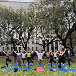 Group outdoor yoga class with a dozen people on colorful mats practicing tree pose under sprawling oak trees in a city park in front of a stone civic building — lively bachelorette/wellness vibe.