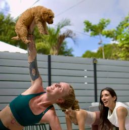 Woman in a teal sports bra holding a small fluffy tan puppy overhead while smiling friends watch in a fenced backyard patio with palm trees