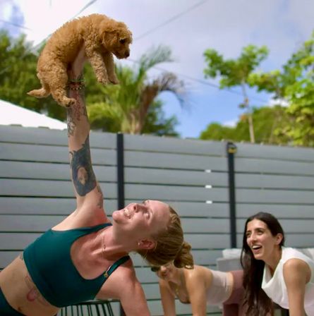Woman in a teal sports bra holding a small fluffy tan puppy overhead while smiling friends watch in a fenced backyard patio with palm trees