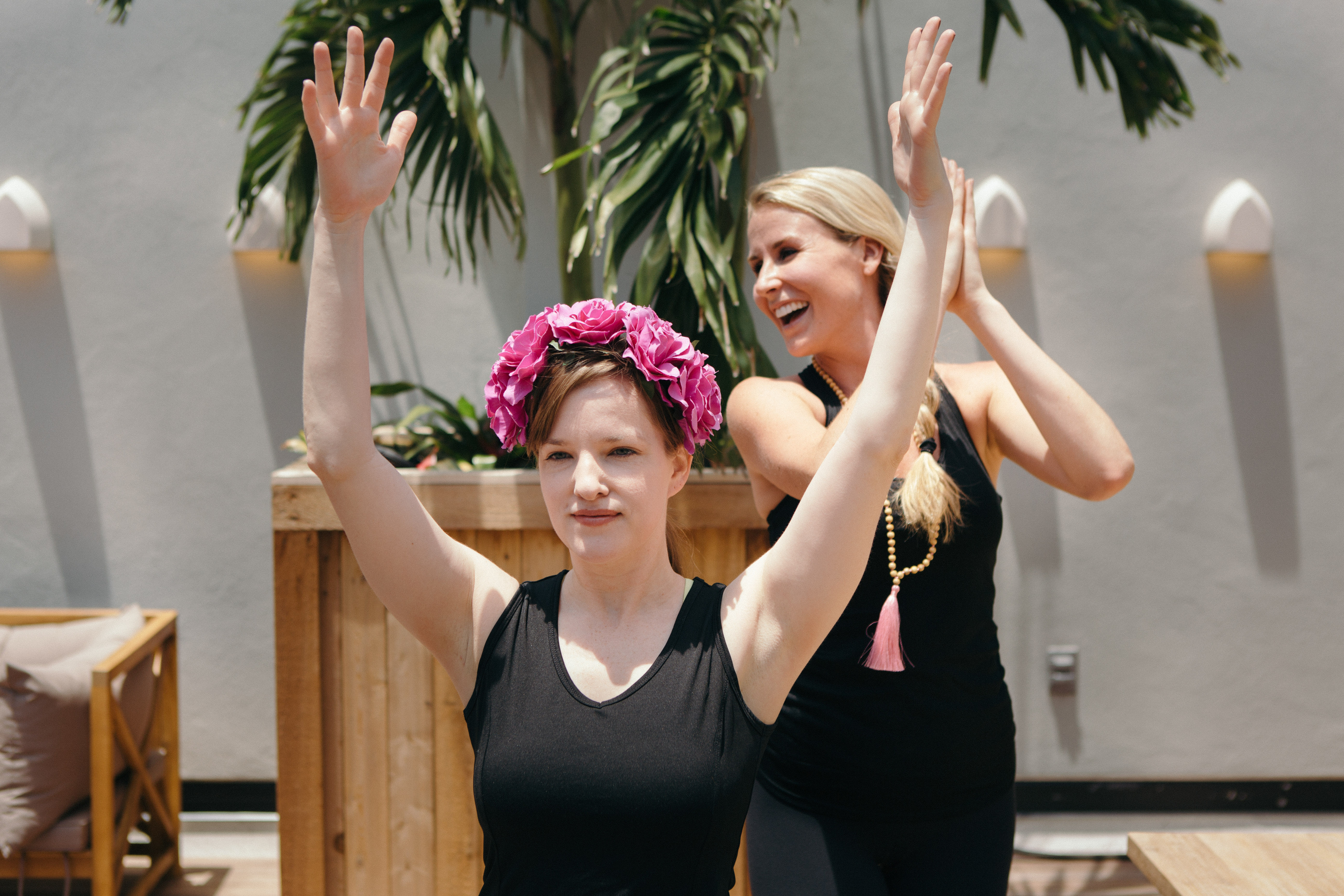 Two women in black workout clothes on a sunny rooftop patio doing yoga — front participant wearing a bright pink flower crown raises her arms while an instructor behind claps and smiles near potted palm plants.