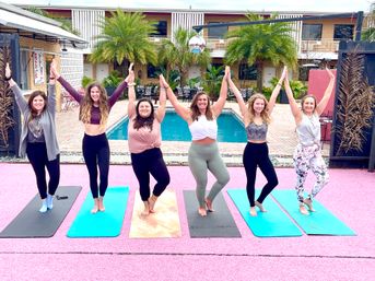 Six smiling women in athletic wear striking tree pose on colorful yoga mats by a palm-lined poolside courtyard during an outdoor yoga class.