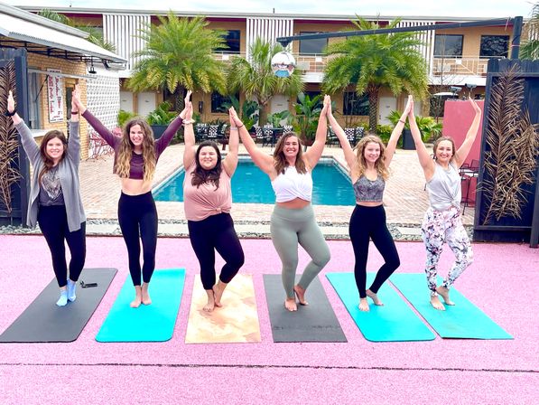 Six smiling women in athletic wear striking tree pose on colorful yoga mats by a palm-lined poolside courtyard during an outdoor yoga class.