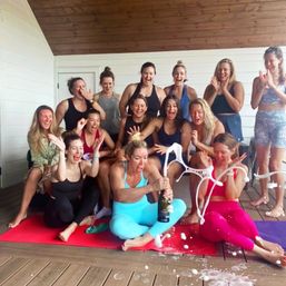 Group of women on a covered wooden deck seated on colorful yoga mats, popping a champagne bottle that sprays foam as everyone laughs and celebrates