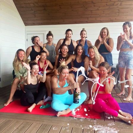 Group of women on a covered wooden deck seated on colorful yoga mats, popping a champagne bottle that sprays foam as everyone laughs and celebrates