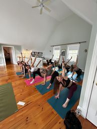 Energetic group yoga class in a bright upstairs loft: people on colorful mats doing side-angle poses on polished wooden floors beneath a vaulted ceiling and ceiling fan, light streaming through windows.