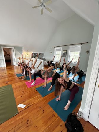 Energetic group yoga class in a bright upstairs loft: people on colorful mats doing side-angle poses on polished wooden floors beneath a vaulted ceiling and ceiling fan, light streaming through windows.