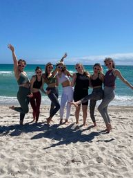 Seven women in activewear and heart-shaped sunglasses posing barefoot on a sunny sandy beach with turquoise ocean waves, arms raised and kicking playfully.