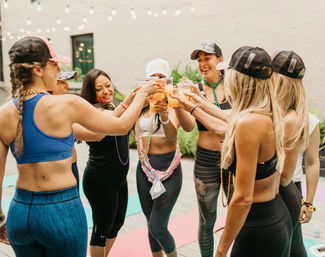 Group of women in athletic wear and caps toasting with plastic cups of orange juice at an outdoor patio yoga/fitness gathering under string lights.