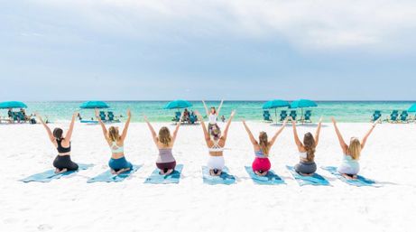 Seven women practicing group yoga on blue mats on a white-sand beach facing the turquoise ocean, arms raised toward the sky with teal umbrellas and lounge chairs in the background.