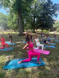Outdoor yoga class on a sunny park lawn under large trees — a group of women on colorful mats doing lunge stretches, foreground participant in bright pink activewear.