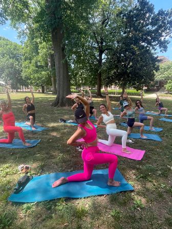 Outdoor yoga class on a sunny park lawn under large trees — a group of women on colorful mats doing lunge stretches, foreground participant in bright pink activewear.