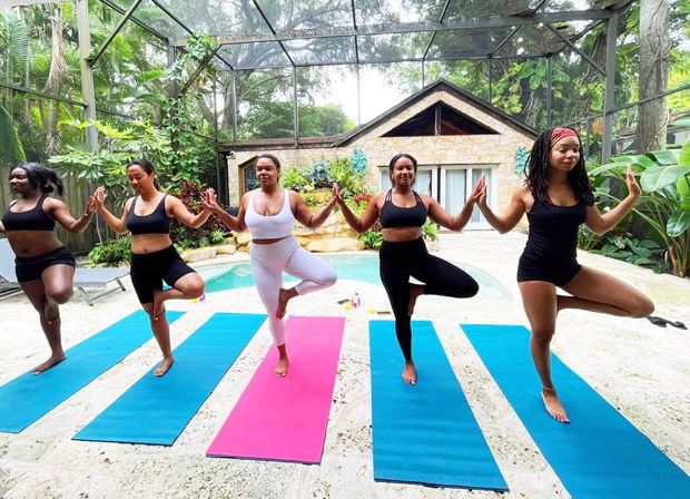 Five women doing tree pose on colorful yoga mats in a poolside outdoor yoga session inside a screened tropical backyard patio.