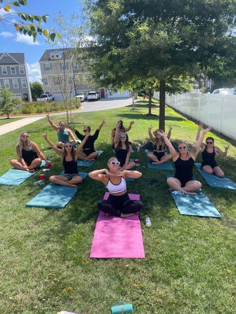 Sunny outdoor yoga session on a suburban lawn: a group of women on colorful mats under a tree, smiling and raising arms during a casual group fitness class near townhouses and a white fence.