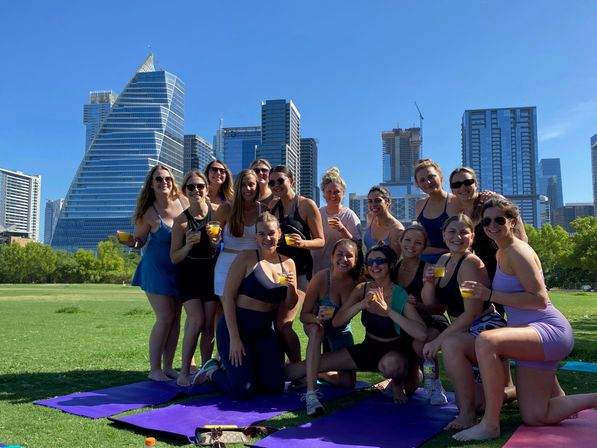 Group of women in activewear holding orange drinks on purple yoga mats during a sunny outdoor gathering in a city park with modern downtown skyscrapers and blue sky