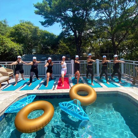 Group of women doing outdoor yoga on a sunny backyard pool deck in warrior pose on mats, with gold inflatable rings and blue floats in the clear pool and trees in the background.