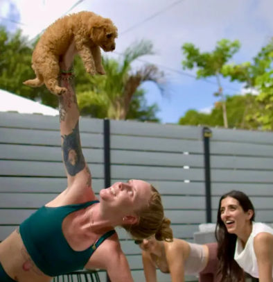 Woman in teal sports bra holding a small curly brown puppy overhead during an outdoor yoga session in a sunny, lush backyard while other women laugh and stretch nearby.