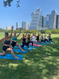 Group of women doing yoga lunges on colorful mats in a sunny urban park, raising arms toward a modern downtown skyline with glass skyscrapers and a distinctive triangular high‑rise.