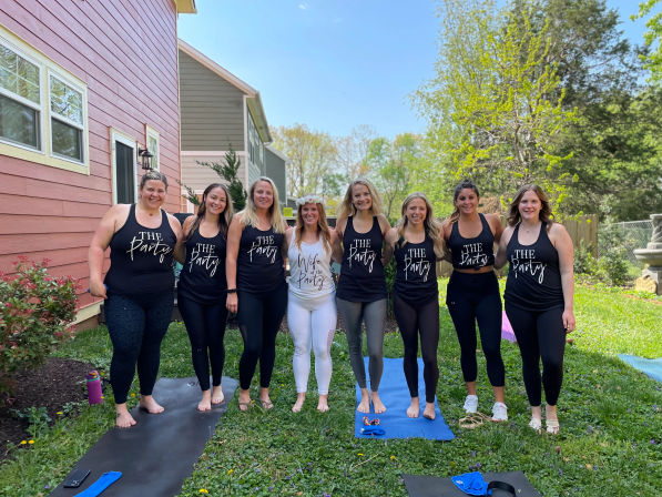 Nine women posing on yoga mats in a sunny suburban backyard; eight wear black tanks reading "The Party" and the center woman wears white "Wife of the Party" with a flower crown for a bachelorette celebration.