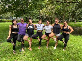 Seven friends in activewear linked arm-in-arm, balancing on one leg on grass in a shaded park with large oak trees, Spanish moss and a fountain in the background — playful outdoor group pose.