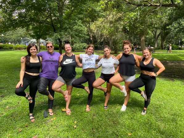 Seven friends in activewear linked arm-in-arm, balancing on one leg on grass in a shaded park with large oak trees, Spanish moss and a fountain in the background — playful outdoor group pose.