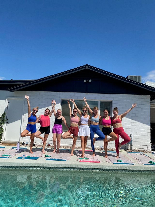 Poolside yoga class with eight women in colorful activewear holding tree pose on mats and towels beside a backyard pool under a clear blue sky