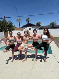 Five friends in sports bras and leggings doing tree-pose arm-in-arm on a pool deck in a sunny suburban backyard with palm trees, string lights, and a clear blue sky.