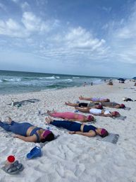 Group relaxation on a white-sand beach: participants lie on towels with eye masks for a seaside yoga/meditation session, ocean waves and blue sky in the background.