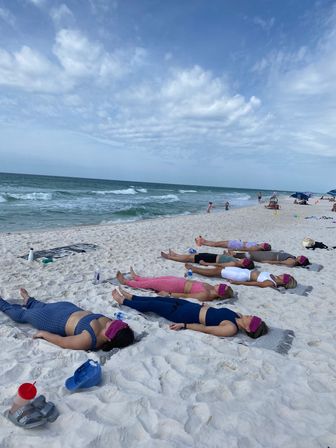 Group relaxation on a white-sand beach: participants lie on towels with eye masks for a seaside yoga/meditation session, ocean waves and blue sky in the background.
