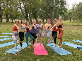 Eight women smiling in tree pose on colorful yoga mats during an outdoor yoga class in a tree-lined park with fallen leaves on the grass