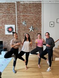 Four friends in an urban loft striking tree pose while holding glasses of orange drink in front of an exposed brick wall decorated with metal stars — bright, casual brunch-yoga vibe.