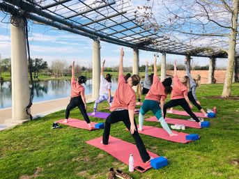 Group outdoor yoga class on colorful mats under a pergola beside a park pond, participants stretching in a warrior pose on a sunny day